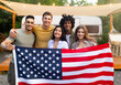 © Prostock-studio - Diverse millennial friends holding American flag and smiling at camera in front of motorhome at campsite. Diverse young people celebrating national summer US holiday outdoors