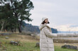 © SHOTPRIME STUDIO - Woman in a light puffer coat and beanie stands outdoors by a lake, calm landscape, cool weather, contemplative mood under an open sky and gentle breeze.