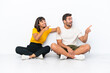 © luismolinero - Young couple sitting on the floor isolated on white background pointing to the side to present a product
