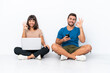 © luismolinero - Young couple sitting on the floor holding pc and mobile phone isolated on white background showing an ok sign with fingers