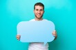 © luismolinero - Young caucasian man isolated on blue background holding an empty speech bubble