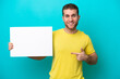 © luismolinero - Young caucasian man isolated on blue background holding an empty placard with happy expression and pointing it