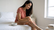 © OlgaNeuroArt - Young woman applying moisturizing lotion to her leg in bedroom. Skin care routine and body wellness concept. Female sitting on bed with cream