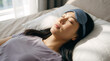 © OlgaNeuroArt - Young Asian woman sleeping in bed with blue eye mask. Relaxed female resting on pillow in morning sunlight. Sleep and wellness concept
