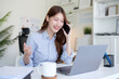 © wattana - Smiling businesswoman talking on a smartphone while working on a laptop at a bright home office desk.