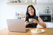 © wattana - Young Asian woman holding a cup of coffee while working on a laptop at home.
