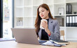 © wattana - Businesswoman drinking coffee while working on a laptop in a modern office.