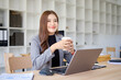 © wattana - Businesswoman drinking coffee while working on a laptop in a modern office.