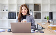 © wattana - Smiling businesswoman holding a coffee cup while working on a laptop in a modern office.