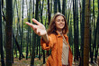 © SHOTPRIME STUDIO - Woman in bamboo forest smiling and reaching hand toward camera, portrait outdoors in nature wearing orange jacket, relaxed expression among tall green bamboo stalks and trees.