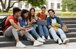 © Prostock-studio - Students Leisure. Group of multiethnic college friends resting in campus outdoors, happy young multicultural men and women using laptop together, relaxing after classes, chatting and laughing