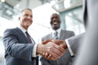 © EricMiguel - Caucasian businessman and Black executive seal a deal with firm handshake — navy suit and purple tie, light - gray suit in bright glass atrium