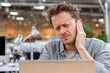 © EricMiguel - Young male office worker wincing and clutching his right ear at a laptop in a bright open - plan workspace wearing a gray shirt