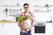 © sofiko14 - An Asian man stands in a modern kitchen holding a paper bag filled with fresh produce
