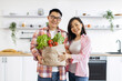 © sofiko14 - An Asian couple smiles while holding a bag of fresh produce in their modern kitchen
