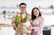 © sofiko14 - An Asian couple smiles while holding a bag of fresh produce in their bright, modern kitchen