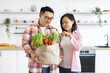 © sofiko14 - An Asian couple smiles while holding a bag of fresh produce in their modern kitchen