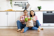 © sofiko14 - A smiling Asian couple sits on the floor with a bag of fresh produce in their modern kitchen