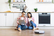 © sofiko14 - An Asian couple sits on the floor in their kitchen, using a laptop while a robot vacuum cleans the floor