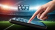 © Margarita Ratatosk - Woman hand touching tablet screen with stadium background. Online sports betting and digital entertainment concept. Modern technology for game.