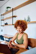 © we.bond.creations - Young woman with curly hair smiling and typing on a laptop, working remotely from her contemporary home office setting