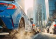 © Daria - Blue sedan among traffic with visible exhaust fumes in a city street during sunset with modern high-rise buildings in the background and warm lighting