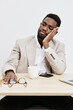 © SHOTPRIME STUDIO - Thoughtful young African American man in beige blazer sitting at desk, resting his head on hand with a pensive expression, surrounded by office supplies, coffee mug, and glasses, isolated on white