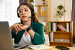 © 220 Selfmade studio - Serious young asian woman wearing headset concentrating on video call looking at laptop screen in home office working online with focused expression