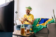 © kleberpicui - Brazilian football fan celebrating at home while watching a match on TV. Adult man wearing national colors, sitting on sofa with snacks and drinks in a casual living room.