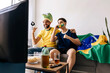 © kleberpicui - Happy father and teenage son celebrating a football moment at home as Brazilian fans. Joyful reaction while watching a match on TV with national colors, snacks and drinks.
