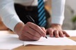 © New Africa - Man writing on sheet of paper with pen at desk indoors, closeup