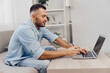 © SHOTPRIME STUDIO - Focused man working on a laptop while sitting on a couch, wearing casual attire, showcasing productivity, modern workspace, and creative vibe.