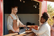 © Seventyfour - Caucasian young adult man processing payment with card reader for Black woman buying strawberries at outdoor market stall, both focused on transaction, digital tablet visible