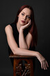 © faestock - close up portrait of beautiful female model with long red hair and black shirt, gestural hand and arm poses, leaning on wooden plinth. classic studio portrait on dark background.