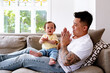 © NZstockphoto - Asian father and baby girl clapping while seated on the couch