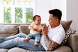 © NZstockphoto - Asian father and baby girl clapping while seated on the couch