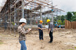 © skarie - Asian female engineer writing on clipboard checklist while male colleagues discuss in background. Quality control manager monitoring building project progress at construction site.