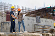 © skarie - Two Asian civil engineers wearing safety helmets looking at blueprint and pointing at building structure. Construction workers supervising progress at industrial site under blue sky.