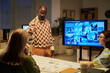 © Mediaphotos - Senior Black man presenting data on large digital screen to two young women seated at table, discussing business charts and graphics during office meeting