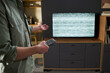 © pressmaster - Midsection shot of man standing in living room holding remote control gesturing toward television displaying static screen, showing frustration with malfunctioning device