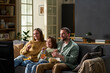© pressmaster - Caucasian young parents and child sitting on sofa watching television together, smiling and eating snacks, enjoying family time indoors