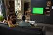 © pressmaster - Caucasian family of parents and child sitting on sofa watching television with green screen in modern living room, backs facing camera