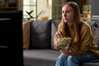 © pressmaster - Caucasian young adult woman sitting on sofa watching television while holding bowl of popcorn, focusing on screen with serious expression, spending leisure time at home