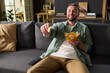© pressmaster - Caucasian young adult man sitting on sofa holding bowl of potato chips and smiling while watching television, extending hand with chip toward screen, remote control on couch