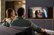 © pressmaster - Caucasian young adult man and Caucasian young adult woman sitting on sofa watching television together in modern living room, both seen from behind with focus on TV screen