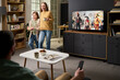 © pressmaster - Caucasian woman and child smiling and walking in living room, while man sitting on sofa holding remote control and watching television with costumes on screen
