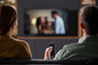 © pressmaster - Caucasian young adult woman and Caucasian young adult man sitting on sofa watching television together, man holding remote control, viewing screen with blurred couple