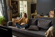 © pressmaster - Caucasian young adult woman sitting cross legged on sofa watching television and holding bowl of popcorn in living room, smiling and focusing on screen