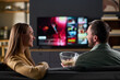 © pressmaster - Caucasian young adult woman and Caucasian young adult man sitting on sofa watching television together, man holding bowl of popcorn, both smiling and engaging in conversation
