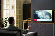 © pressmaster - Caucasian man sitting on sofa watching television news holding remote control in living room, focusing on screen with breaking news headline visible on TV